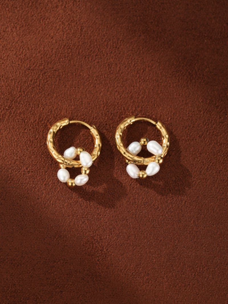 Boucles d'oreilles goutte créoles texturées ton or présentant des grappes de petites perles blanches irrégulières d'eau douce et de perles dorées contre un fond texturé marron.