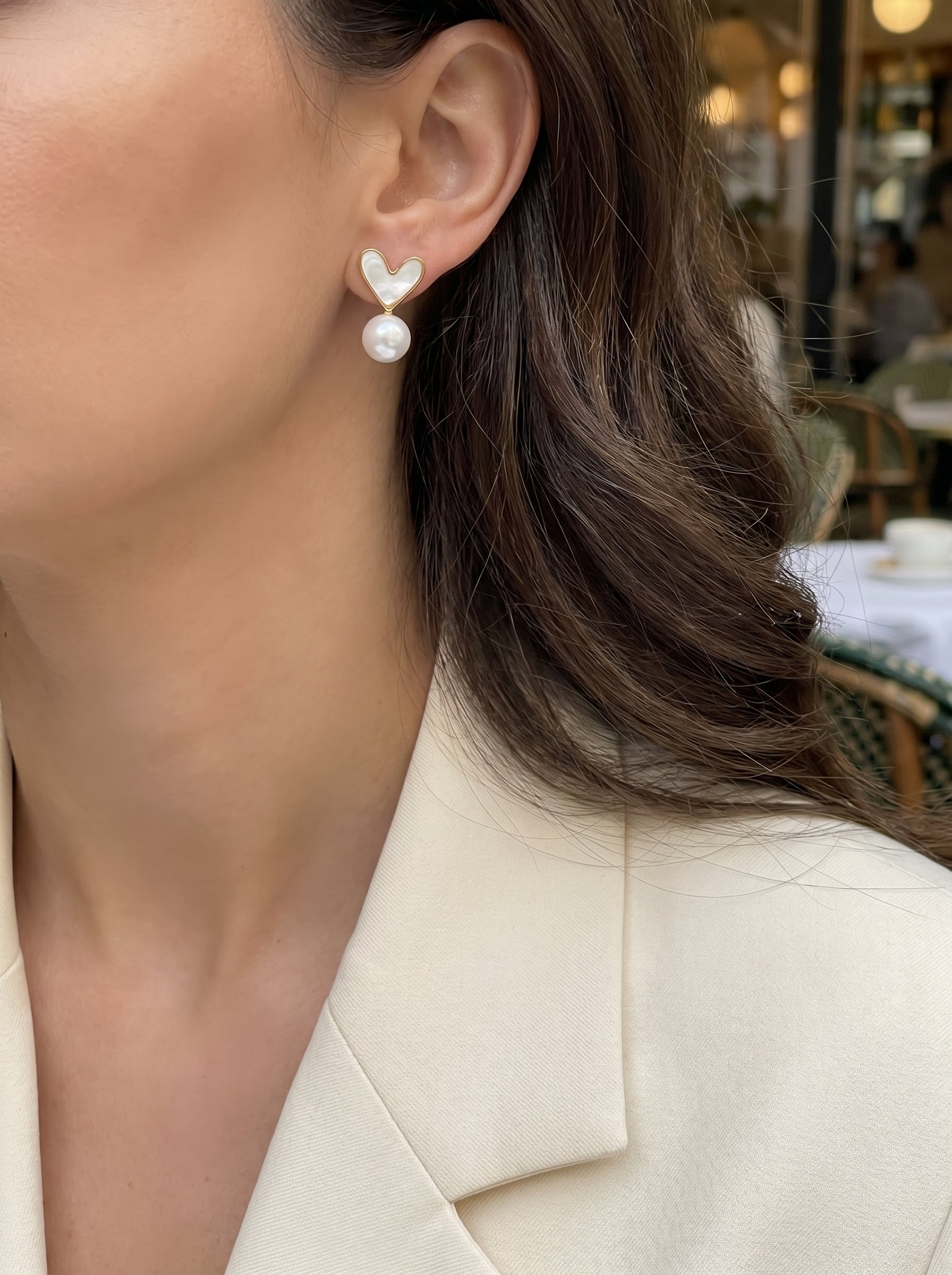Vista - Boucles d'oreilles goutte cœur en perle d'eau douce avec un motif cœur blanc et une perle bouton blanche d'eau douce de 6 mm suspendue, portées par une femme en blazer crème dans un café en extérieur. @lifestyle