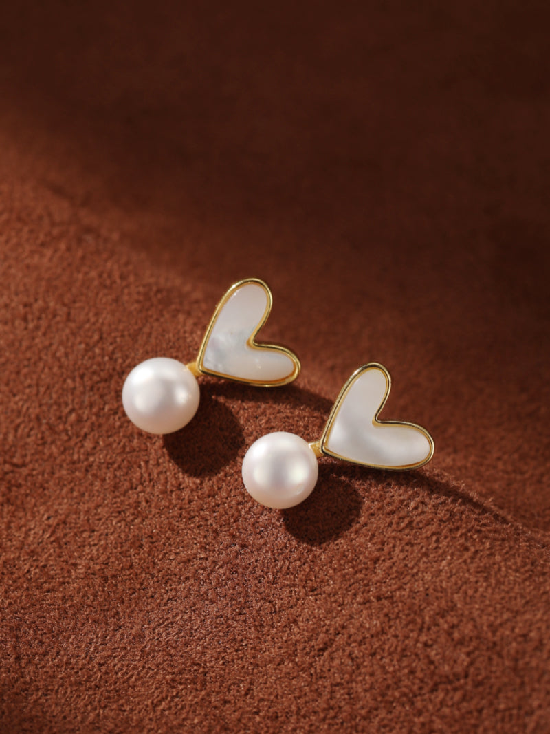 Vista Freshwater Pearl Heart Drop Earrings featuring a delicate white heart charm accented by a lustrous white pearl drop. These earrings are showcased against a textured brown background, highlighting their elegant design.@pearl