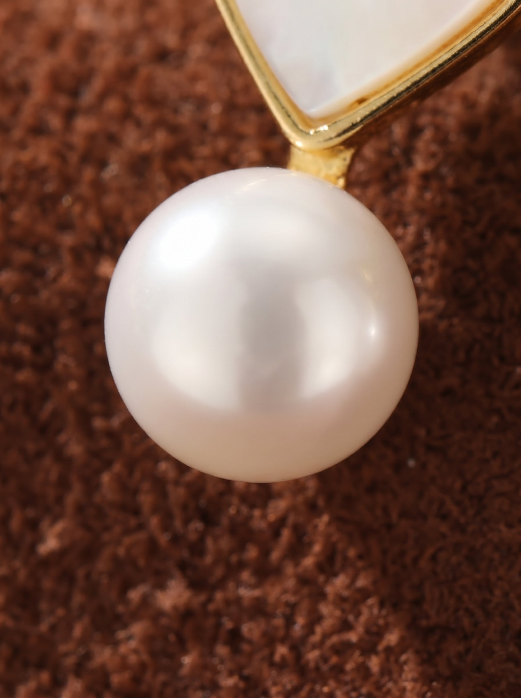 Close-up of a white freshwater pearl drop earring with a gold heart-shaped accent against a textured brown background. @pearl