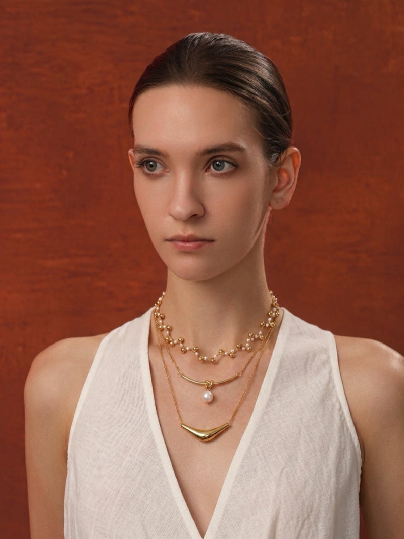 Model wearing the Voyage Freshwater Pearl Knot Layered Necklace, featuring three strands with small pearls and golden beads, a central pearl suspended from a knot, and a sculptural gold-tone bar. @model
