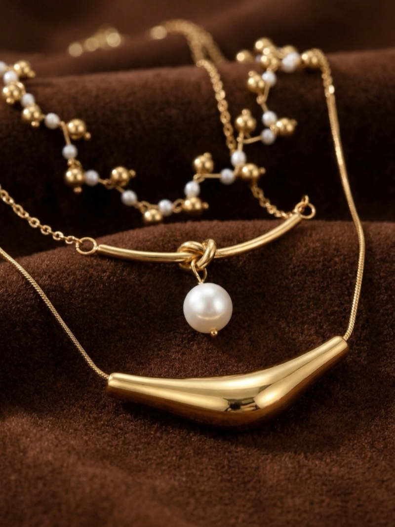 Gold-tone layered necklace featuring delicate freshwater pearls and golden beads on the top strand, a single pearl suspended from a knot detail on the middle strand, and a sculptural bar on the bottom strand.