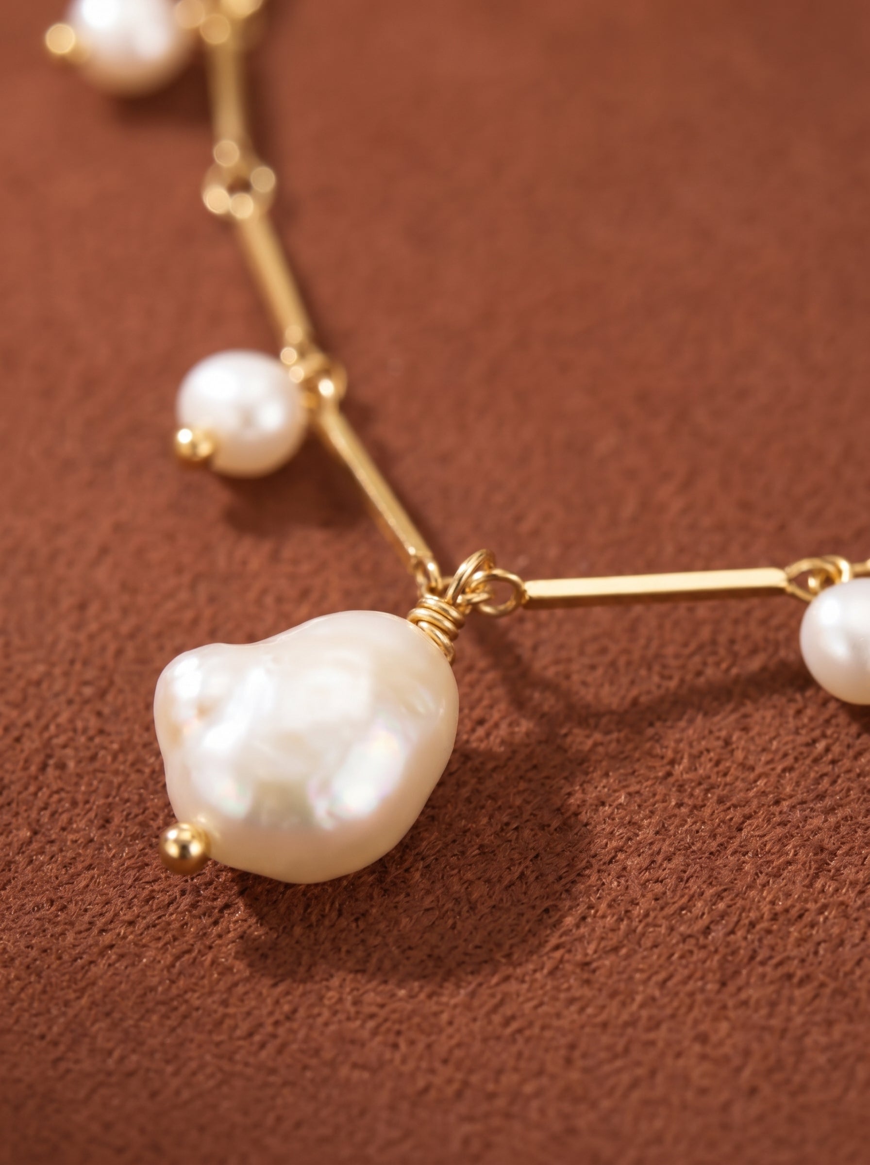 Close-up of a dainty gold chain necklace with multiple luminous freshwater pearls, including a large baroque pearl centerpiece, against a brown textured background. @pearl