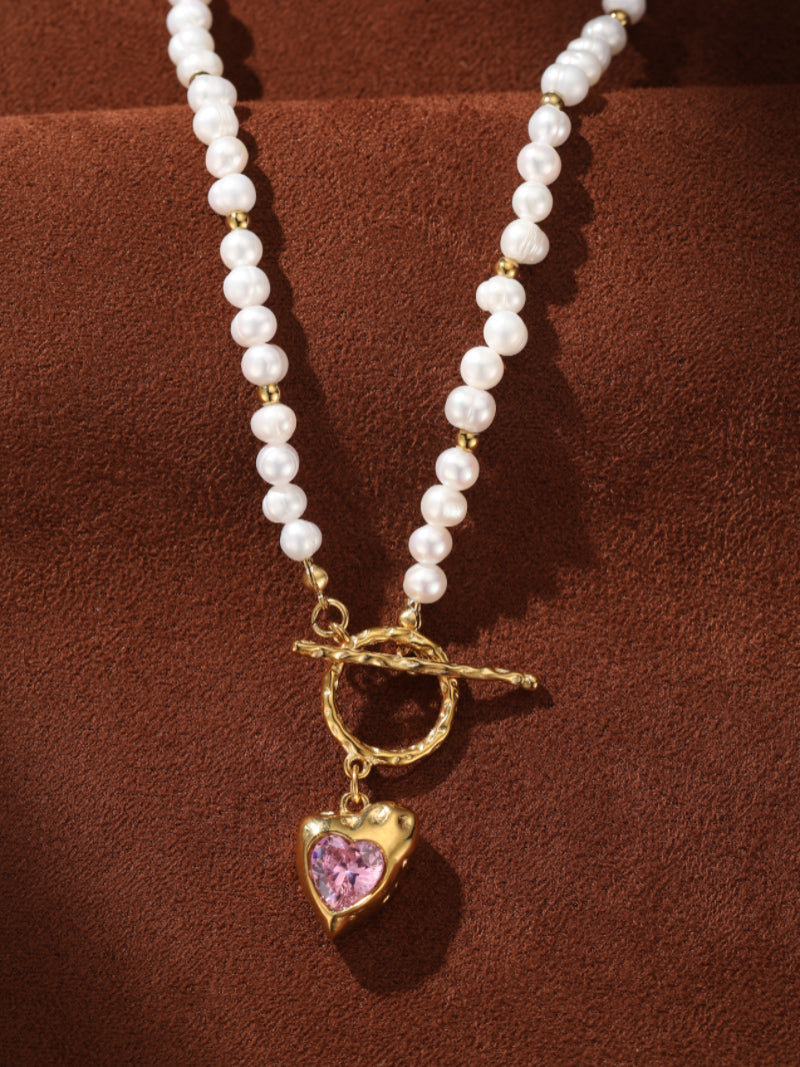 A close-up of a white freshwater pearl beaded choker necklace with gold accents and a gold heart pendant featuring a pink gemstone and a toggle clasp.