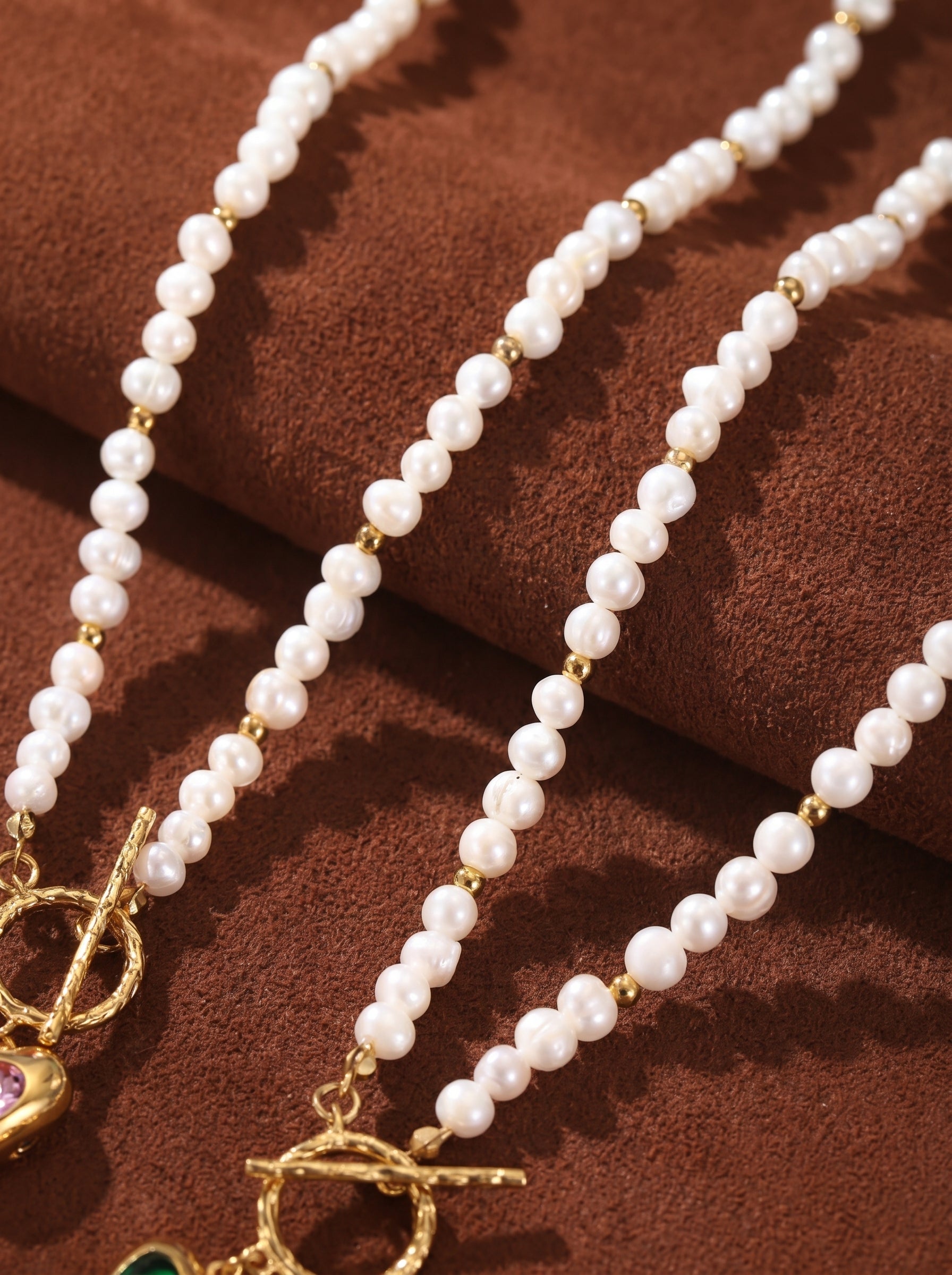 Close-up of two Whimsy freshwater pearl choker necklaces with gold bead accents and heart-shaped pendants on a brown textured background.