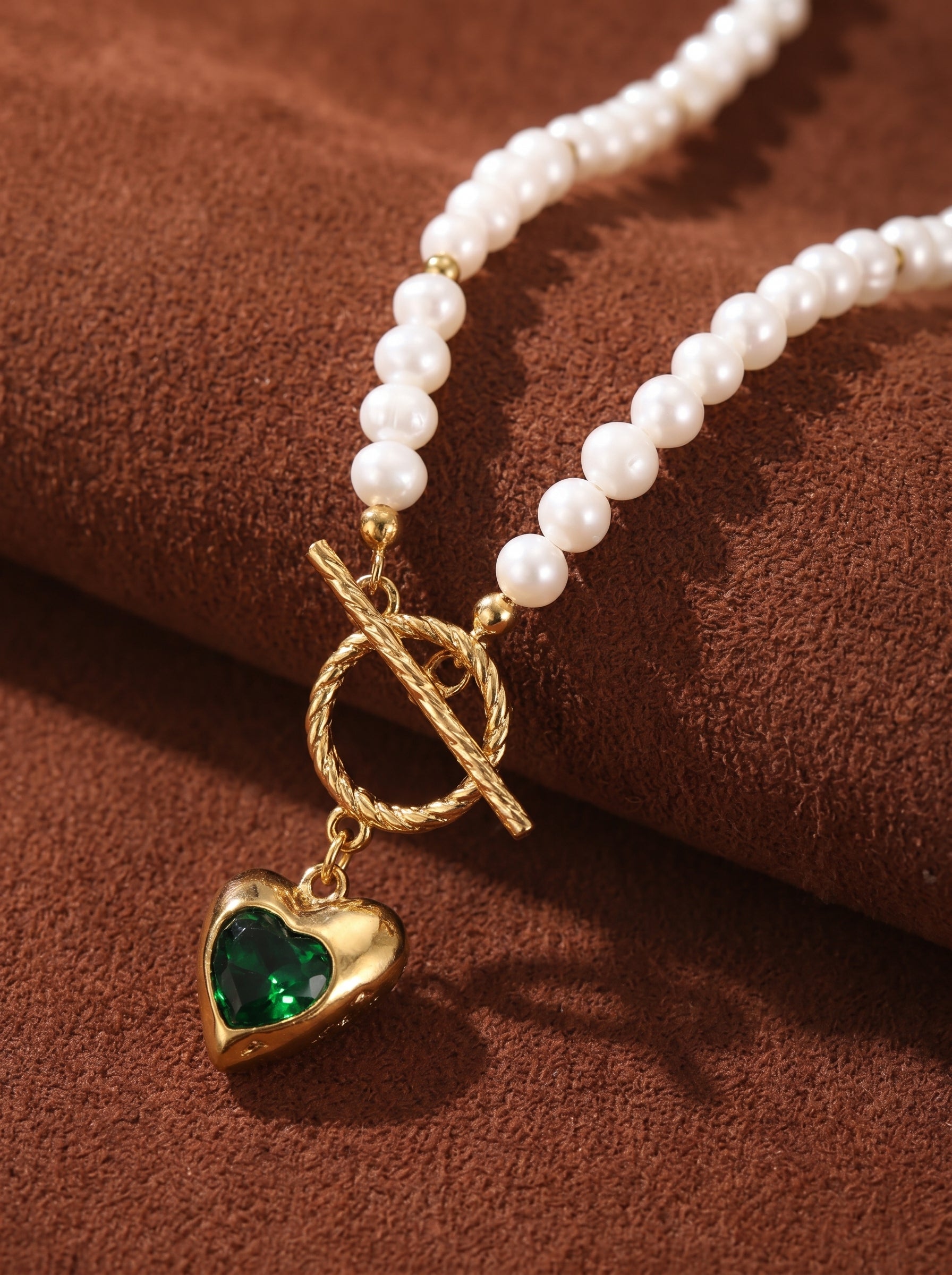 Close-up of a white freshwater pearl choker necklace with gold accents, featuring a gold heart pendant with a green gemstone and a toggle clasp. @pearl
