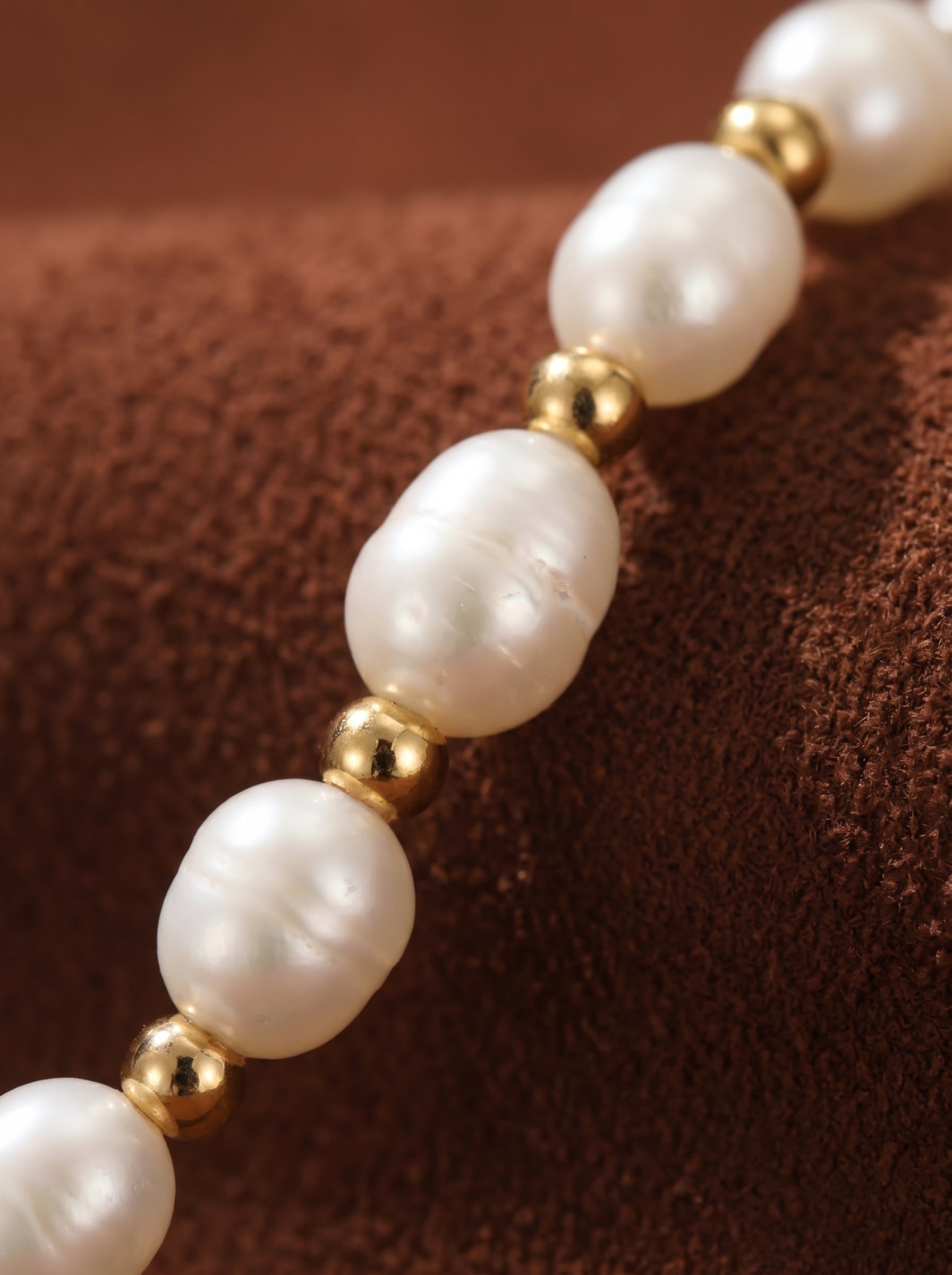 Close-up of the Whisper necklace featuring a strand of lustrous 6mm seed-shaped freshwater pearls interspersed with small, shiny gold beads on a brown textured background. @pearl