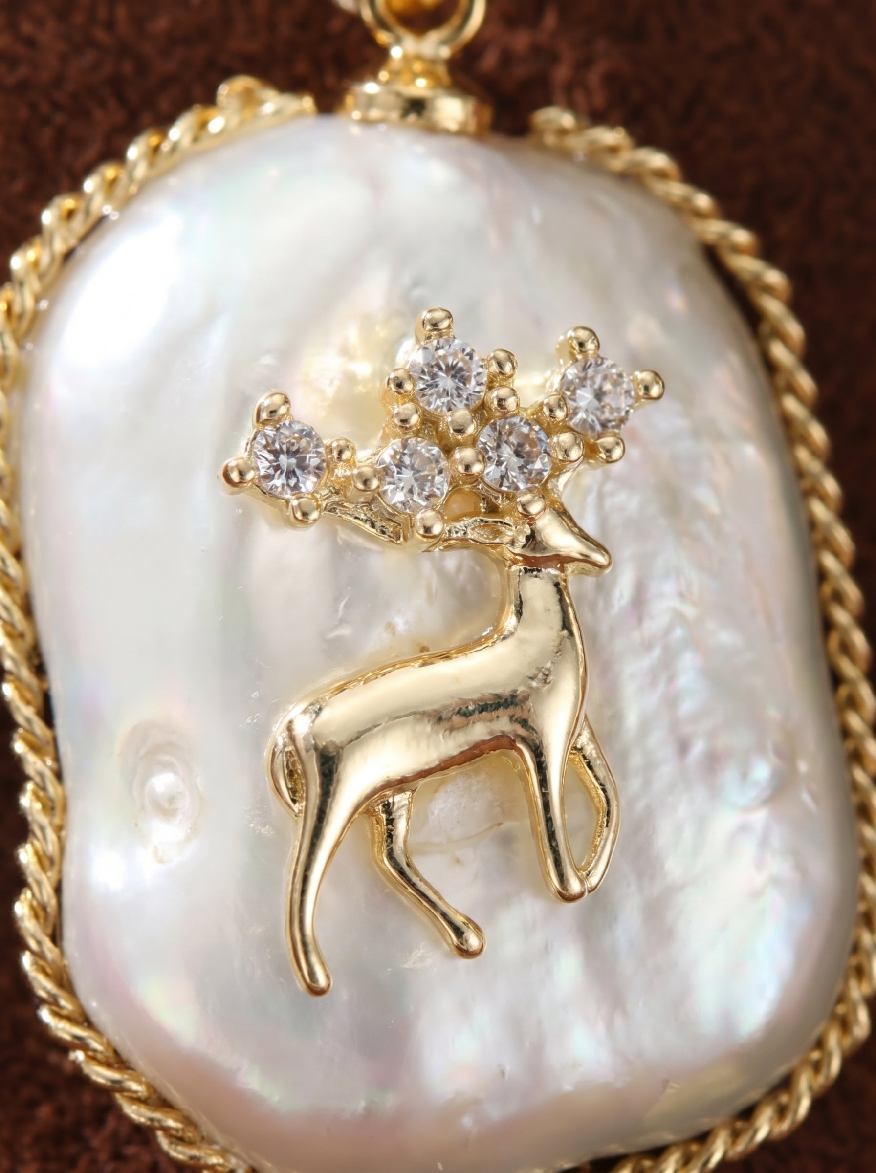 A close-up of a single Wild Grace earring featuring a lustrous baroque freshwater pearl with an iridescent white sheen, encircled by a golden rope-like border. A detailed golden deer motif with sparkling crystal antlers stands on the pearl. @pearl